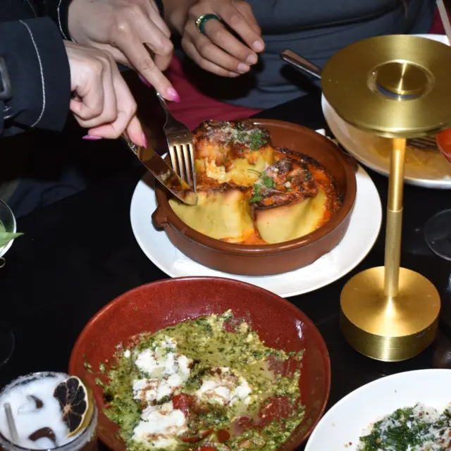 Overhead shot of a shared restaurant meal with baked pasta, a pesto and burrata dish, and colorful cocktails on the table