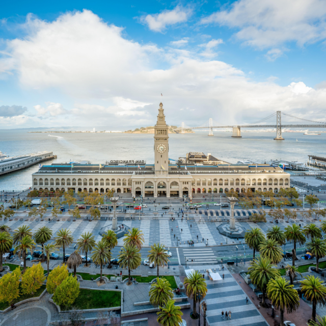 Aerial view of San Francisco's Ferry Building and clock tower, with palm-lined plaza in front and the Bay Bridge across the water