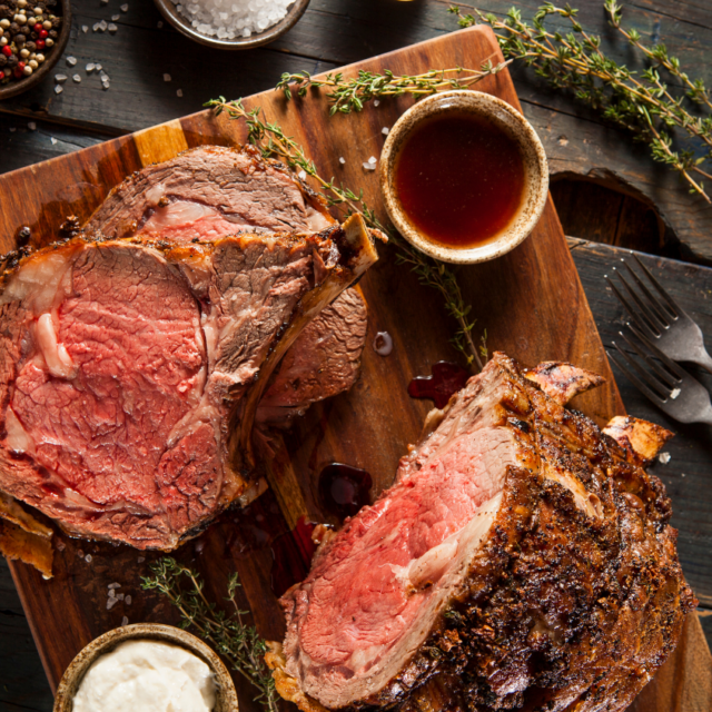 Overhead shot of sliced prime rib roast on a wooden board, surrounded by au jus, herbs, and side dishes.