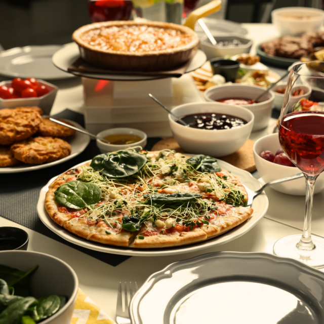 Full spread of dishes on a dinner table, including a leafy flatbread, a pie, and glasses of red wine