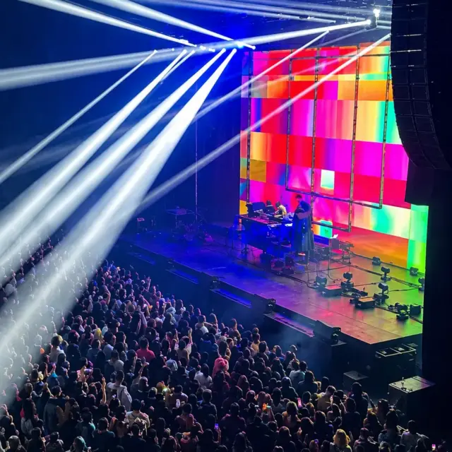 Concert crowd watching a performer on stage, lit by crossing white spotlight beams and a colorful LED backdrop.